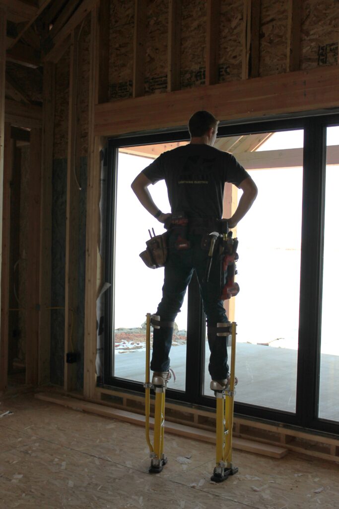 electrician standing on stilts with electrical bags in new construction